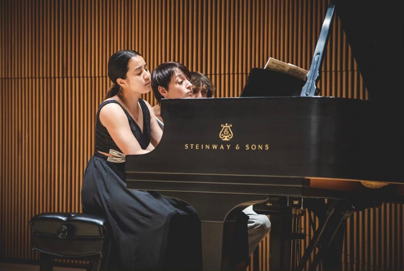 Leah Gavilan in recital at a Steinway, warm wood-panelled hall behind.